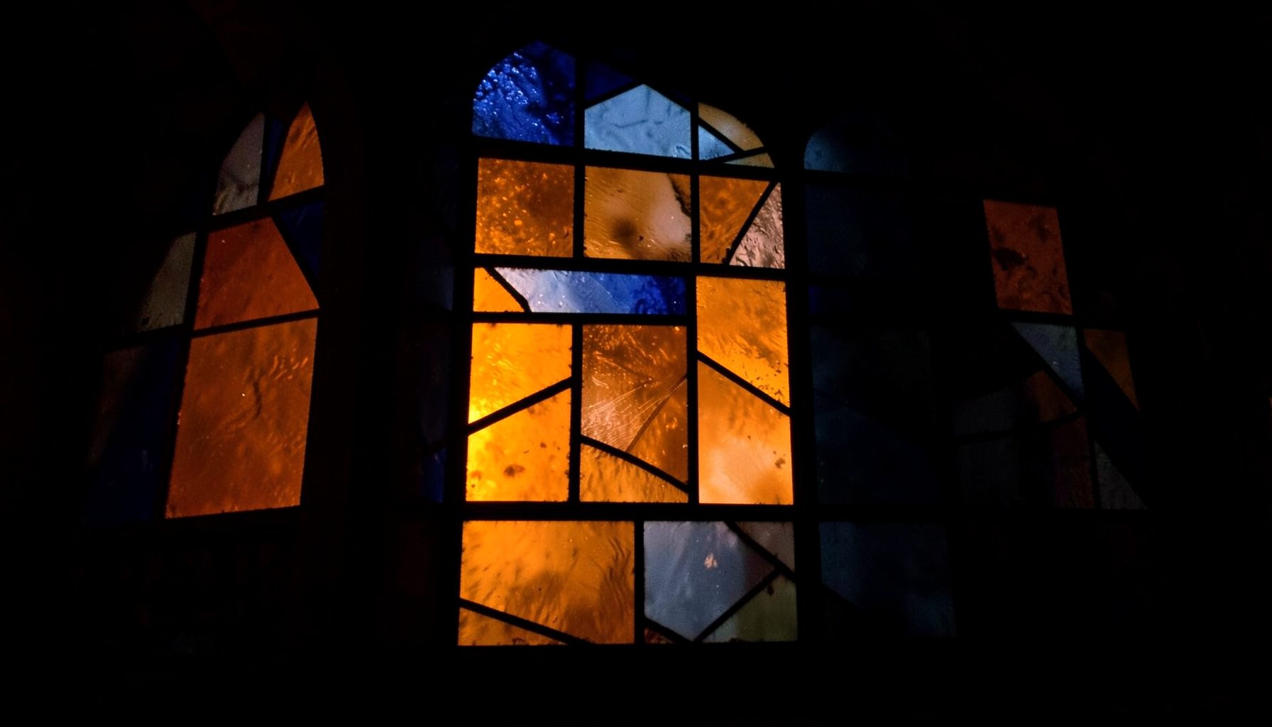Stained-glass composition with bold leaded segments in deep amber, navy, and slate, casting warm geometric color into a moody dark interior